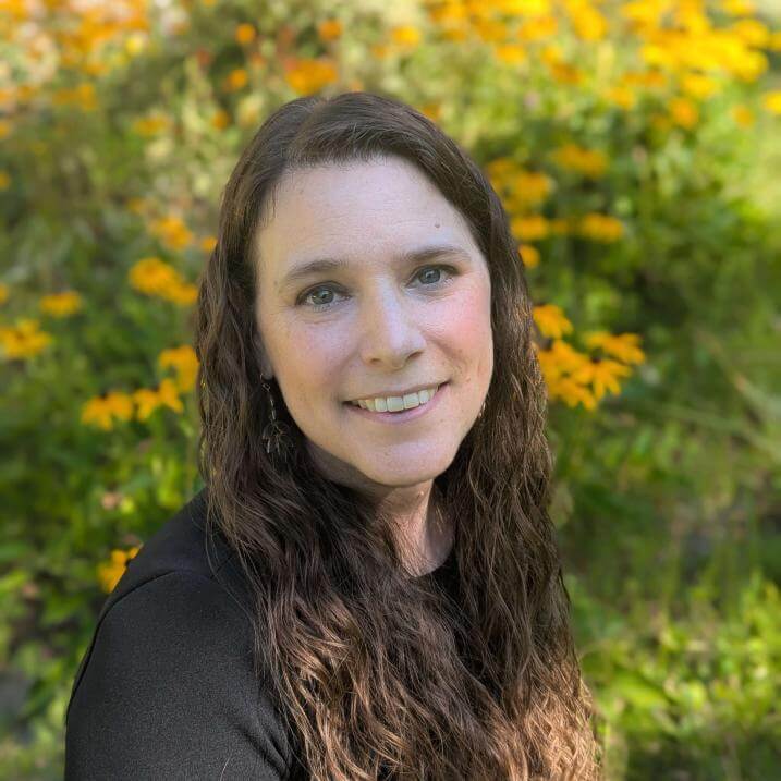 Julie Chiappinelli headshot: pale-skinned woman with long dark wavy hair and dark eyes, looking straight into the camera in a field of tall yellow flowers.