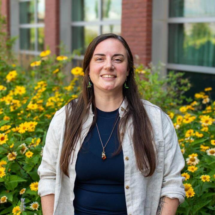 Headshot of Samantha Hoff, she is standing in front of yellow flowers, she has long brown hair and is smiling