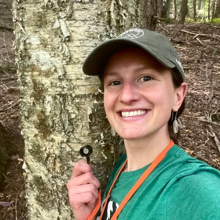 Clara smiles and holds a hand lens in front of a paper birch tree