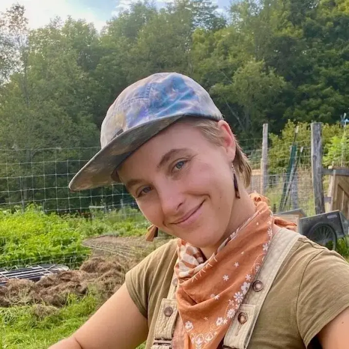 Lena in a blue cap and bandana around her neck, by a garden