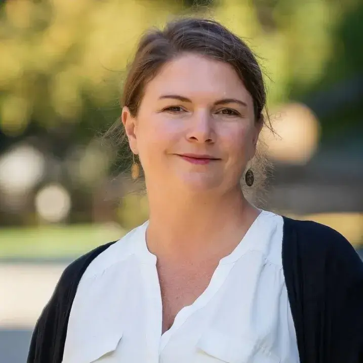 Headshot of Kiersten wearing a white shirt and black house coat. Fall foliage is in the background.