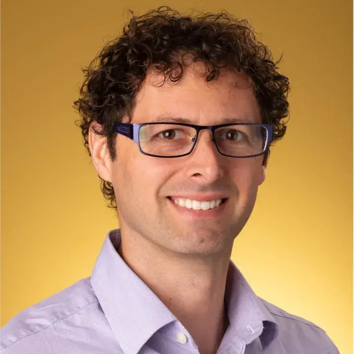 Headshot of smiling man with glasses, purple shirt against a yellow background.