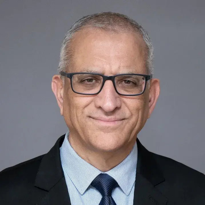 Headshot of a person who has short white and black hair and glasses, wearing a blue striped shirt, dark navy blue tie, and a black sport coat in front of a gray background.