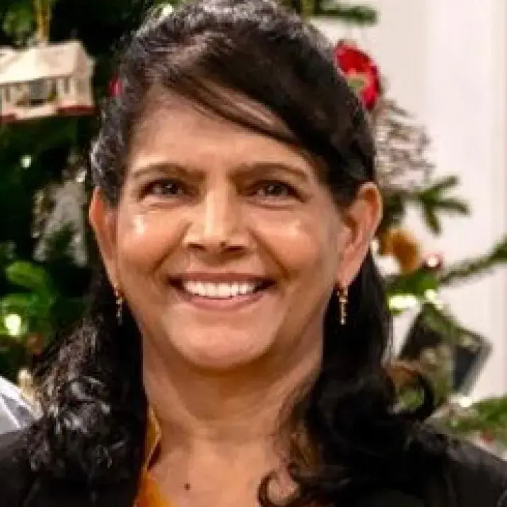 Headshot of an individual with long black hair, wearing earrings, in front of a Christmas tree 