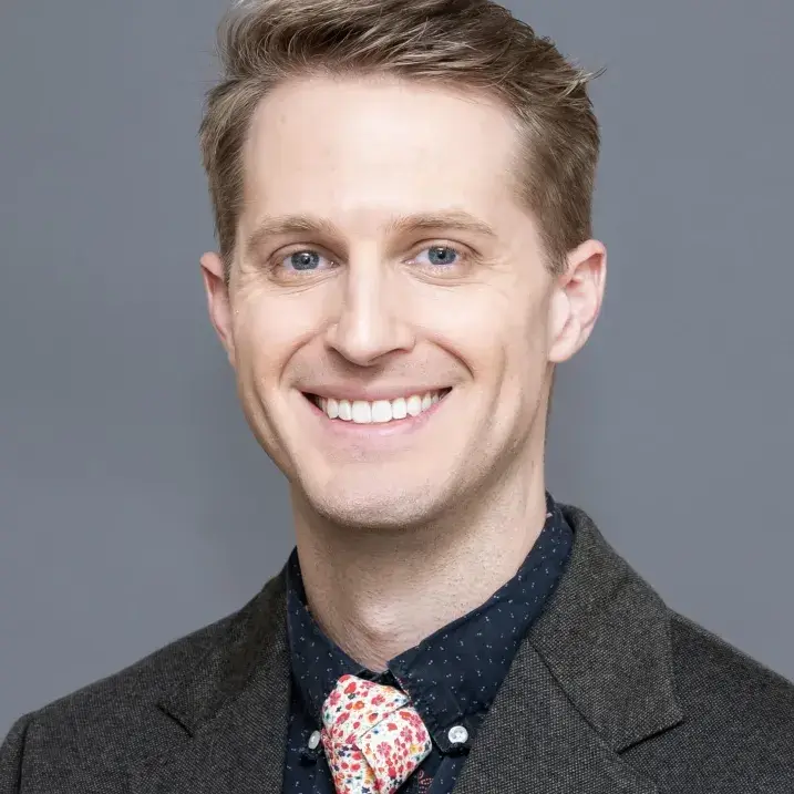Headshot of individual wearing a charcoal gray sport coat, navy blue shirt, and white and pink-flowered tie.  
