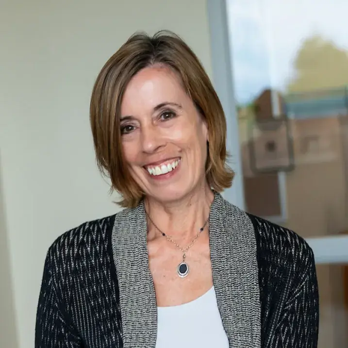 A photo of an individual with medium-length brown hair, wearing a white blouse, black and gray sweater. 