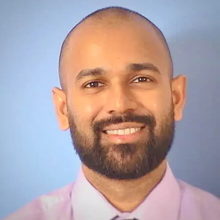 Photo of an individual with closely cut hair, and dark beard, wearing a pink shirt with a tie, standing in front of a light blue background.