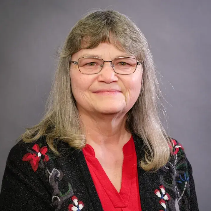 A person with long, light-colored hair wearing a red blouse and a dark cardigan decorated with red and white floral designs, posed against a plain gray background.