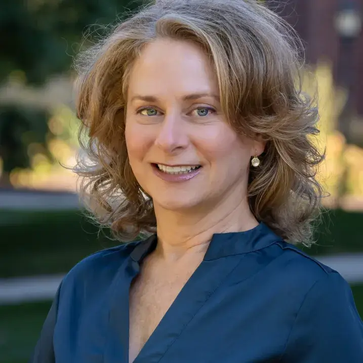 A pale-skinned woman in her mid-forties with lightly curled dark blonde hair, wearing a cobalt blue blouse with mandarin collar. She is smiling slightly at the camera.