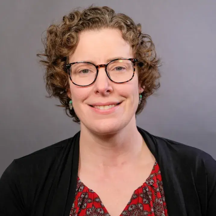 A photo of an individual with curly medium-length auburn colored hair, wearing dark-rimmed glasses, turquoise and silver earrings, and a black cardigan over a red blouse with paisley flower pattern.  