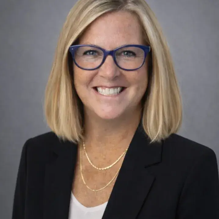 Sharon Mone: pale-skinned woman with a blonde sidepart bob and glasses, in a black blazer and white shell