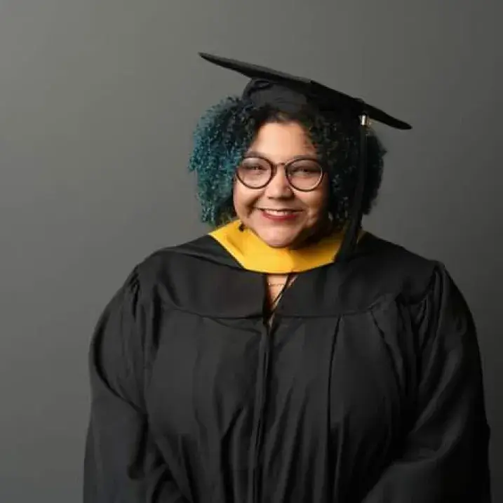 Emily wearing a black graduation cap and gown with a gold hood stands against a plain gray background. They have short, curly hair with teal highlights, are wearing round glasses, and face the camera with a warm, confident expression.