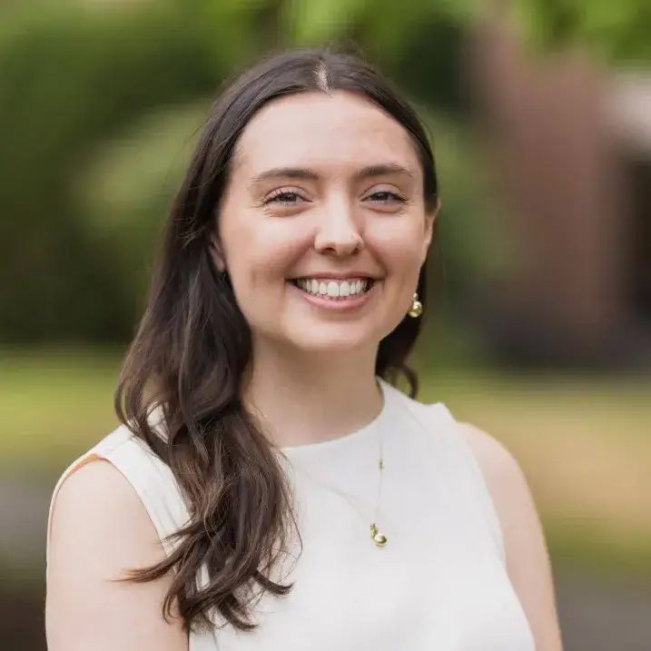 Green tree background, Julia is smiling looking into camera. She is wearing beige top