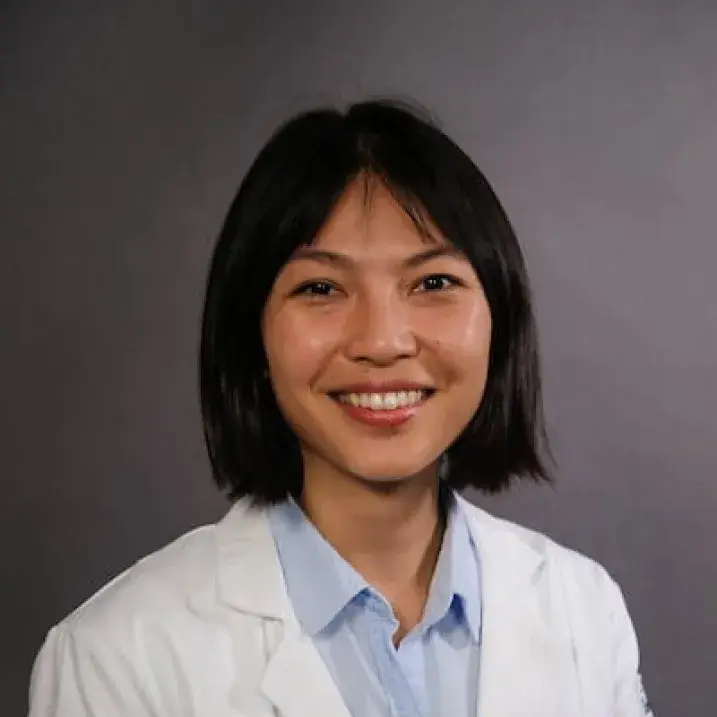 Smiling woman with straight shoulder length black hair, wearing a white doctor's coat and a light blue button down blouse.