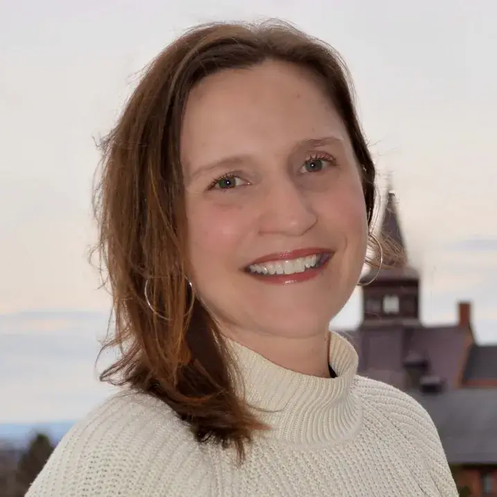 Molly is a white woman with shoulder length brown hair. She is standing outside with the UVM skyline behind her.