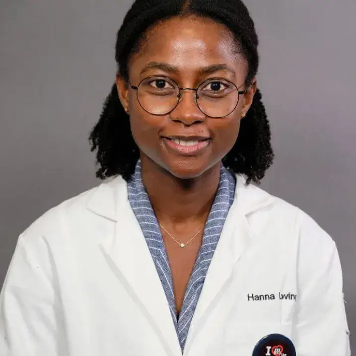 A smiling young African American woman wearing dark-rimmed glasses, a blue blouse with white stripes, and a white doctor's coat.