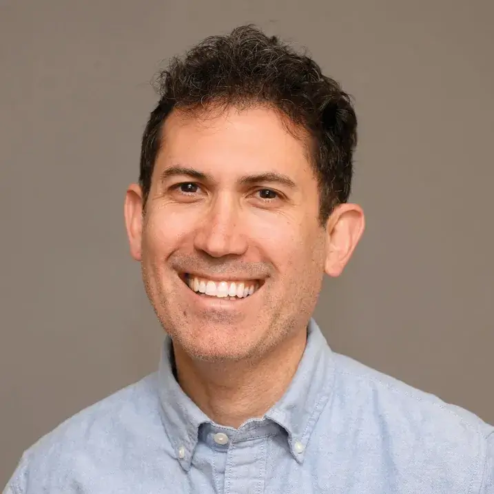 person with a light bear, dark short curly hair wearing a blue button down shirt