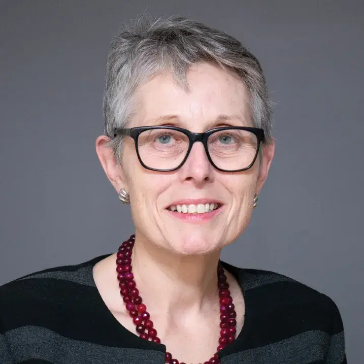person with short grey hair, dark rimmed glasses and a red beaded necklace