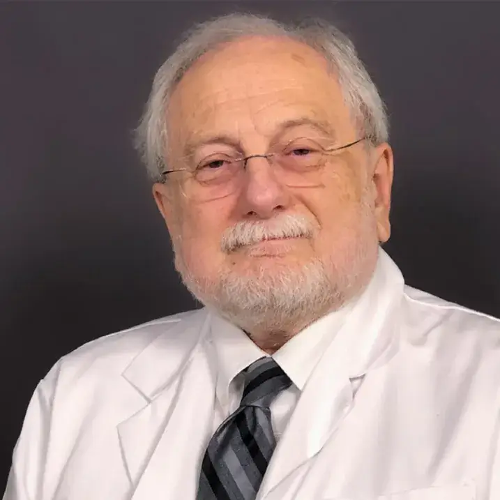 man with light skin, grey hair and beard wearing a striped tie and white doctors coat