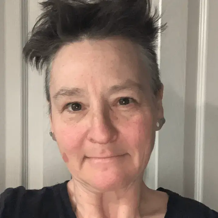 Middle-aged white non-binary person with short dark hair going wild on top, and dark eyes.