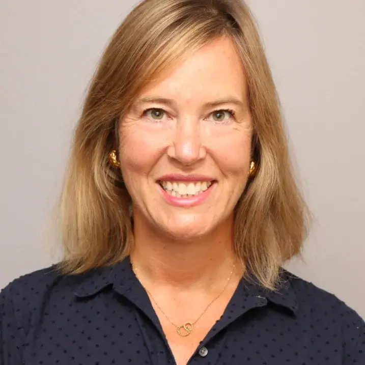 Woman with blonde hair and green eyes smiling. She is wearing a dark navy-blue button-down blouse with raised polka dots.   
