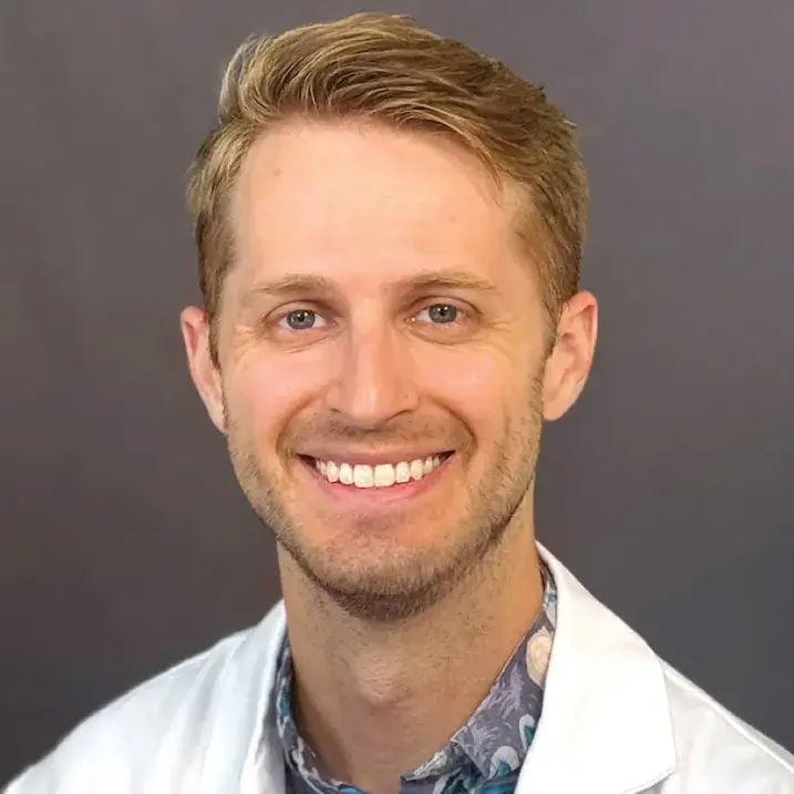 Strawberry blonde male smiling wearing a purple shirt with large white flowers with blue stems and a white doctor's coat.