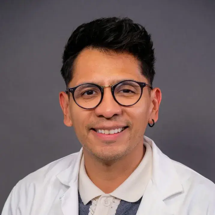 olive complexioned man with short black hair and round rimmed glasses wearing a white doctors coat