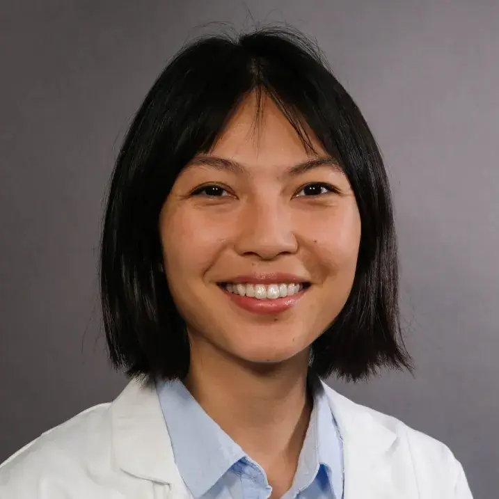 Woman with shoulder length black hair smiling wearing a light blue blouse under a white doctor's coat.