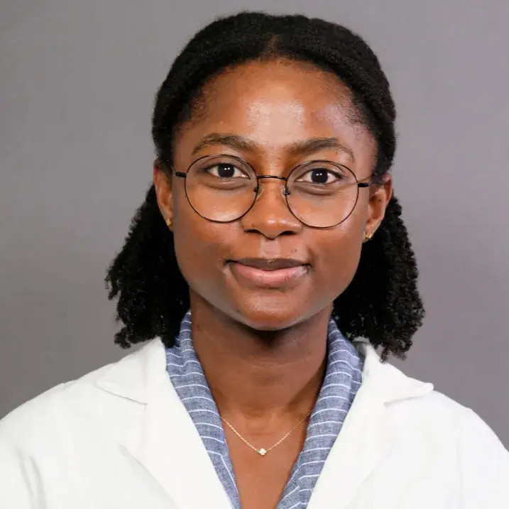 A smiling young African American woman wearing dark-rimmed glasses, a blue blouse with white stripes, and a white doctor's coat.