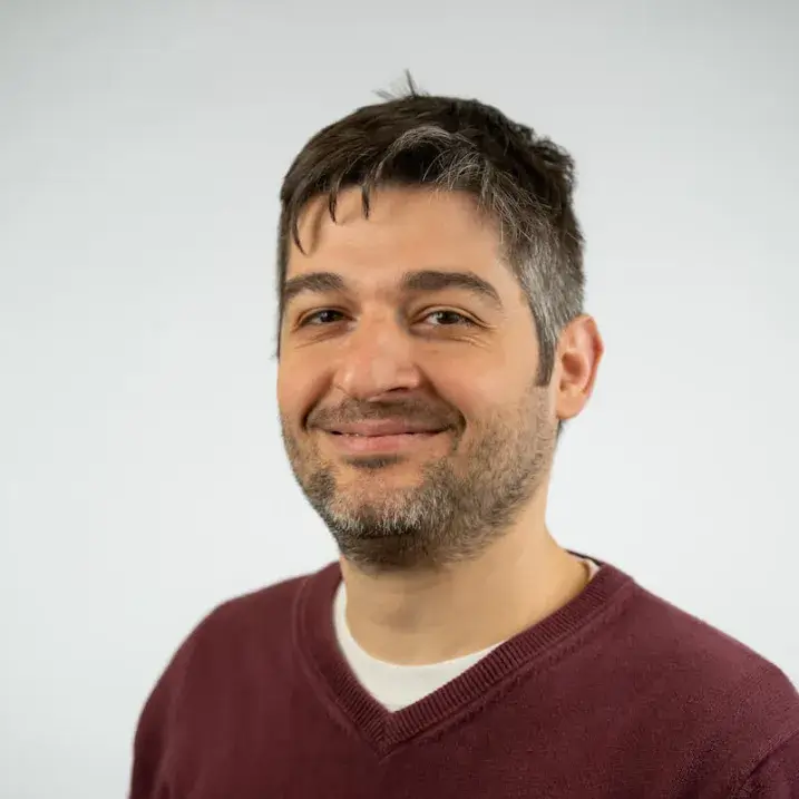 a person with short brown hair and beard wearing a white t-shirt under a maroon v-neck sweater