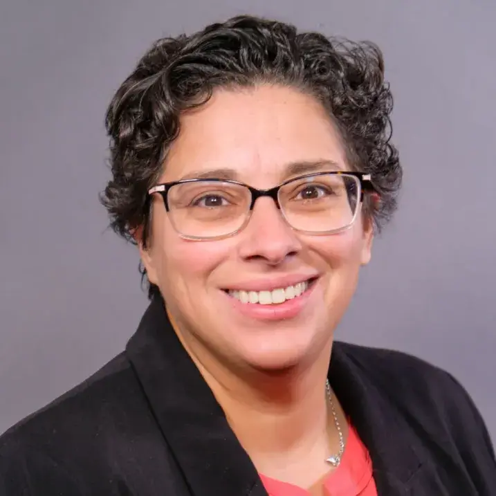 Smiling woman with short curly black hair wearing glasses and a black blazer over a red blouse.