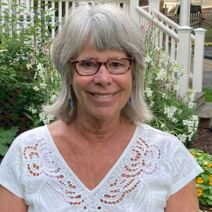 A woman with short gray hair, bangs and glasses wearing a white shirt. 
