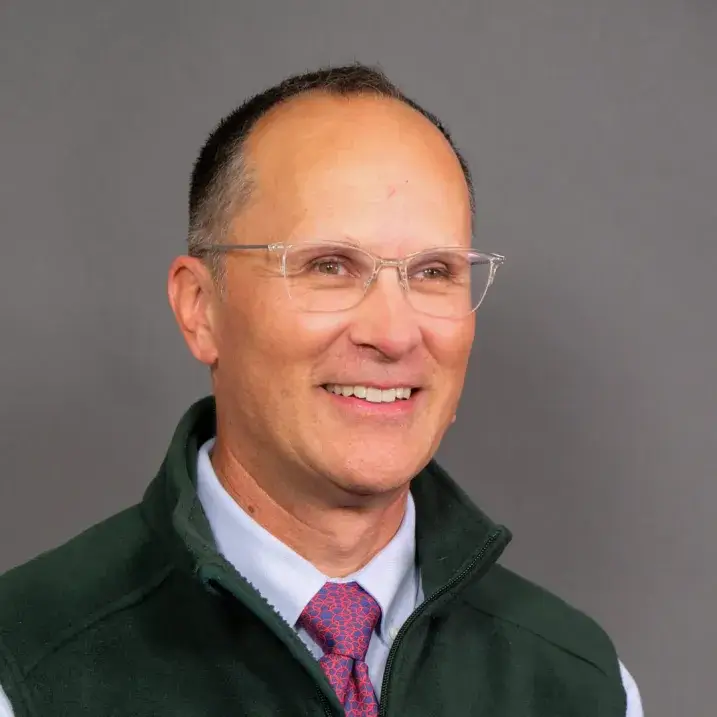 light skinned man with glasses, short gray hair wearing a dress shirt, tie and green fleece vest