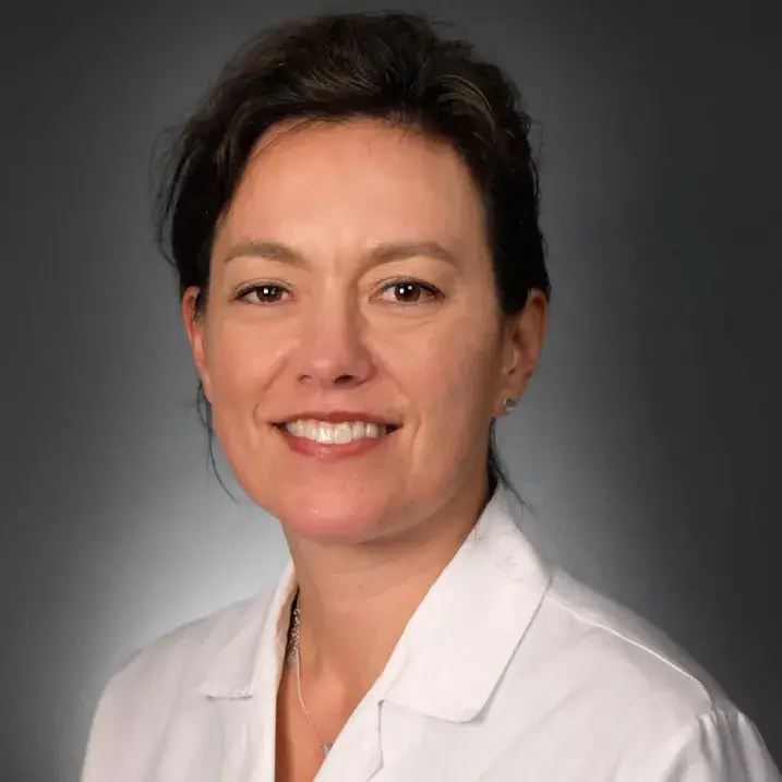 Woman with black hair tied back, smiling, wearing a white doctor's coat.