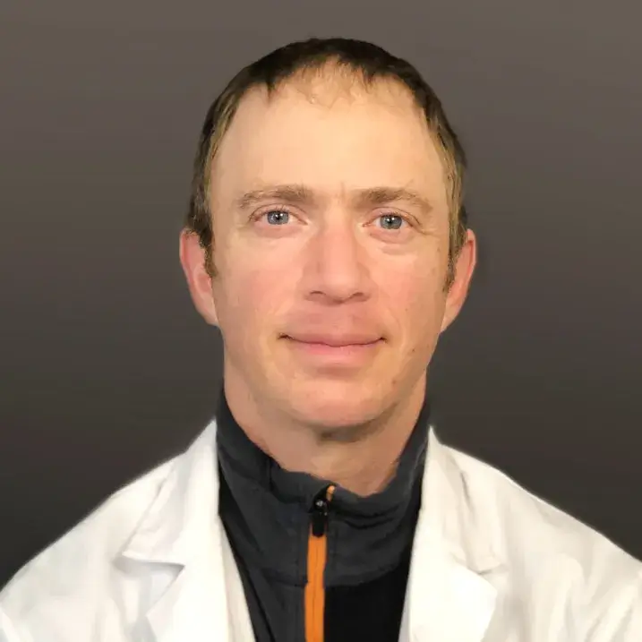 a person with short brown hair wearing a dark green fleece under a white doctors coat