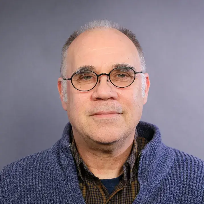 light skinned man with glasses, short gray hair wearing a plaid shirt and blue cardigan