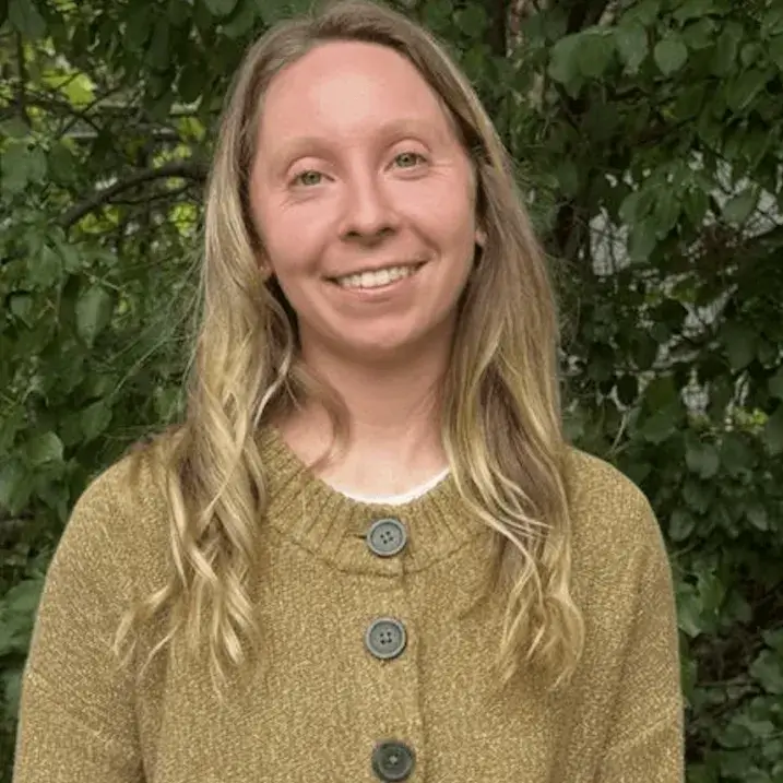 Nicole posing in front of background of green leaves. Nicole is in the center, smiling, and wearing a dark beige cardigan with chunky black buttons. 