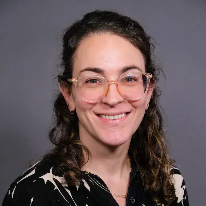 A person with long dark hair and glasses wearing a black patterned blouse