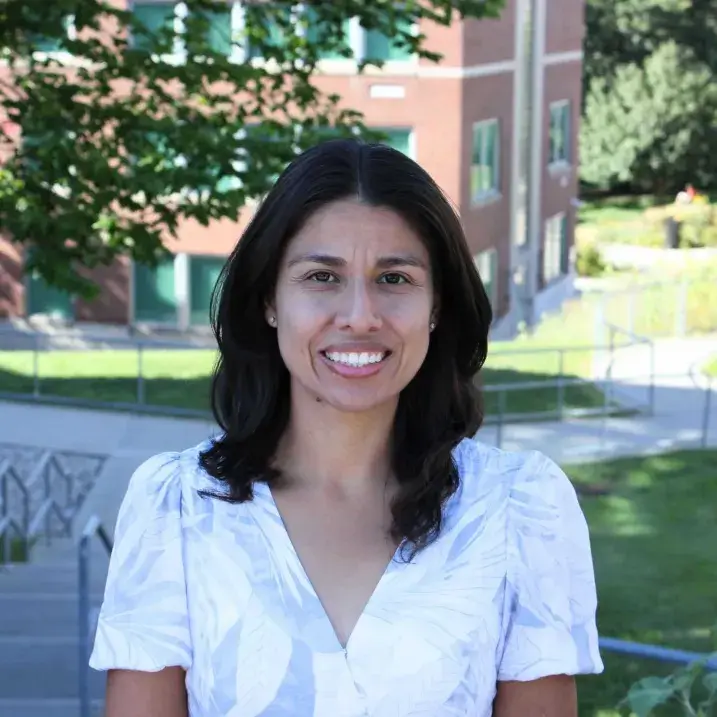 Laura Sanchez-Parkinson smiling in a white shirt with green foliage and a brick building behind her.  