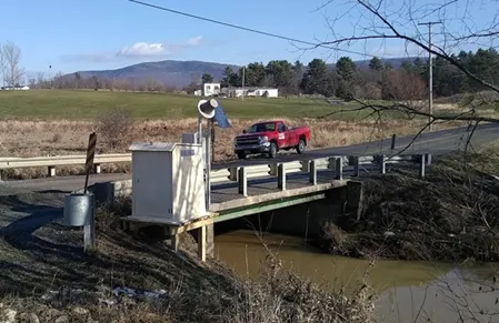 image description: a white box with instruments protruding from it sits on the edge of a roadside over a stream