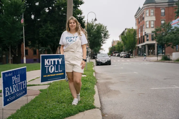 A woman wearing a Kitty Toll for Vermont t-shirt stands in front of a Kitty Toll campaign side staked along a street in downtown Burlington.