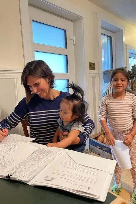 Woman sits at a table completing paperwork while a child sits in her lap. Another child stands in behind her.