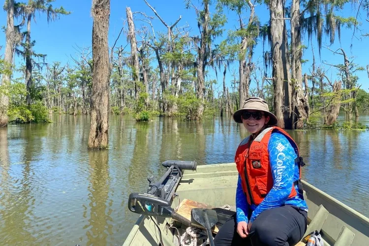 student sitting on a boat in a marsh with large trees growing from the water.