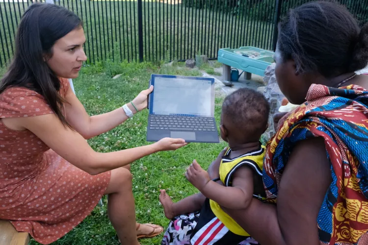 A woman seated on a bench discusses readings on a computer with another woman with two small children sitting on her lap.