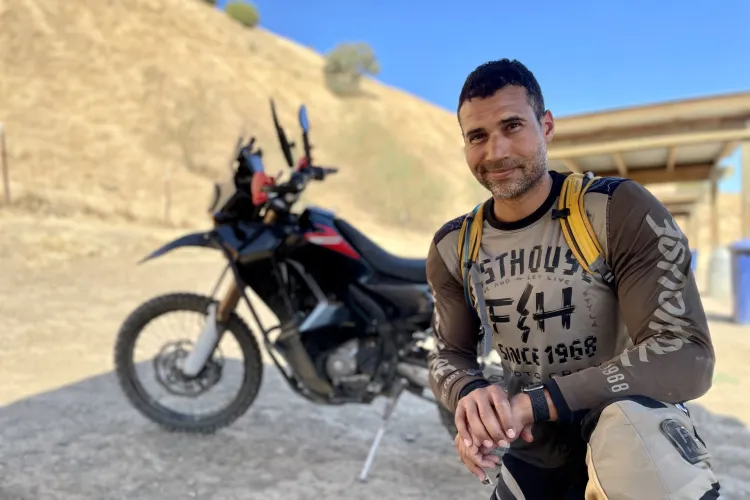 man with dark hair kneeling in front of black and red motorcycle