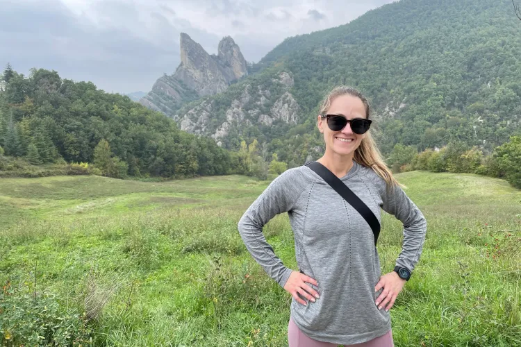 woman with blond ponytail and sunglasses standing in green field with mountains in background