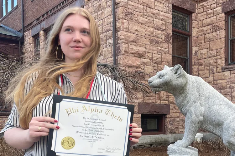 young woman with long blonde hair wearing striped shirt and standing in front of stone building holding Phi Alpha Theta membership certificate