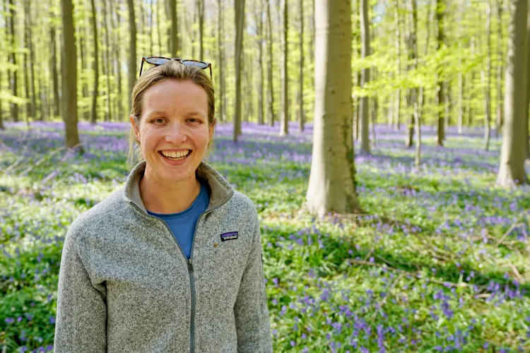 woman with blond hair and gray zip-up sweater standing in sunny forest with purple flowers