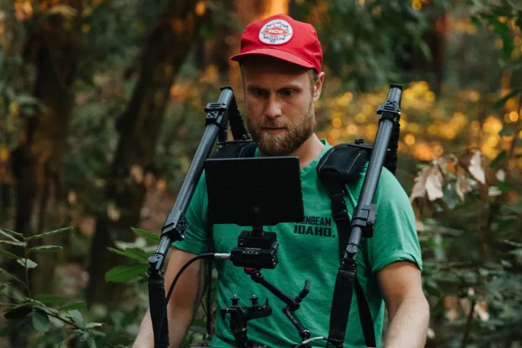 bearded man wearing green t-shirt and red hat shoots a film in the woods