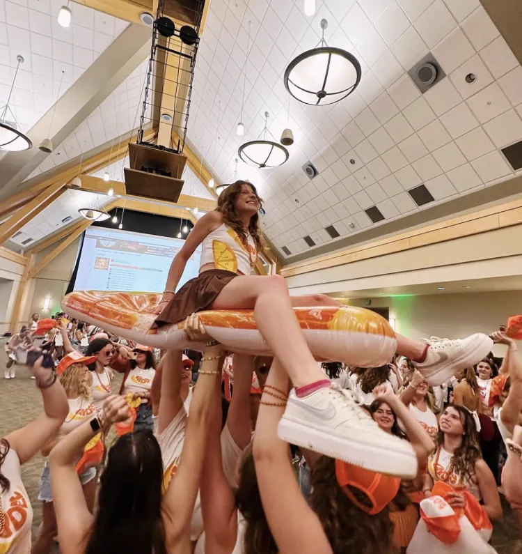 A young white woman with brown hair wearing a white tank top is lifted above the crowd during 2023 Bi Night. Photo by Kappa Delta.